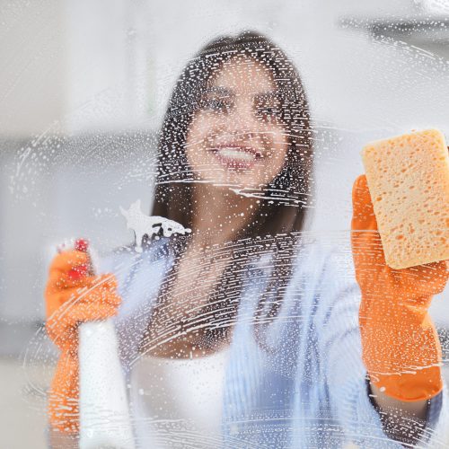 Young woman washing window in rubber gloves with special detergent. Concept of cleaning glass or window at home.