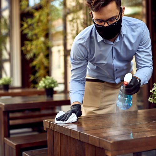 Waiter wearing protective face mask while disinfecting tables at outdoor cafe.